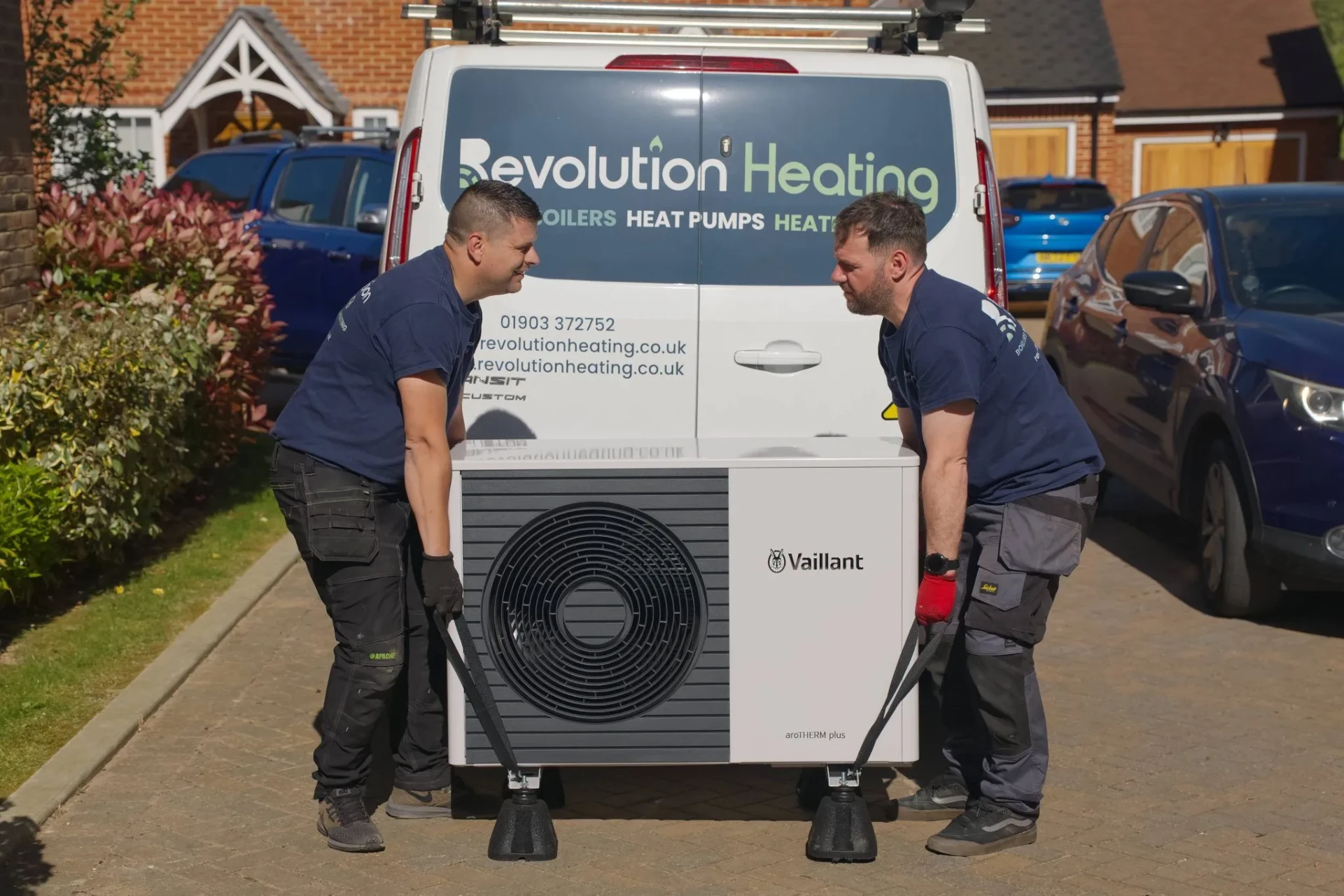 Two Revolution Heating engineers lifting a Vaillant aroTHERM plus heat pump beside the branded van